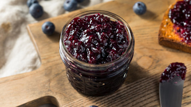 Open mason jar of blueberry jam on wooden board with toast, butter knife, and scattered blueberries