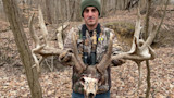 Man in camo jacket and green beanie holding large antlered deer skull in leafless woods