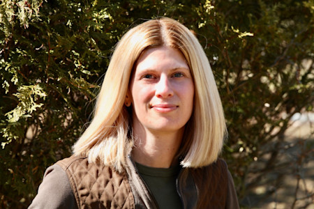 Woman with shoulder-length blonde hair and slight smile wearing brown quilted vest in front of trees