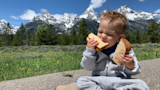 Toddler sitting on curb in front of snowcapped peaks, biting two slices of bread