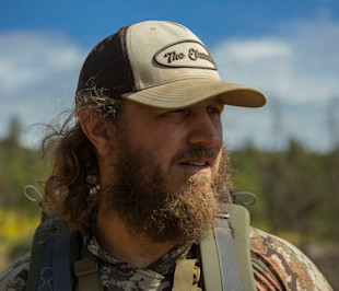 Hunter wearing beige cap reading "The Element", camo jacket and backpack straps, looking right