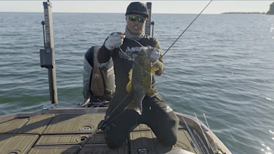 Angler kneeling on boat holding a large bass, wearing sunglasses and cap, open water background