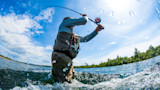 Angler waist-deep in river spey-casting with arched rod and water droplets