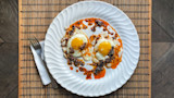 Two fried eggs drizzled with chili crisp on white plate, fork on napkin on bamboo placemat