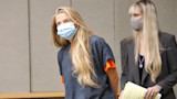 Blonde woman in gray jail uniform and blue surgical mask, hands behind back in courtroom; masked woman behind holds legal papers