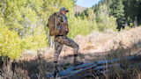 Hunter in camouflage looking back carrying large backpack standing on logs in sunlit meadow