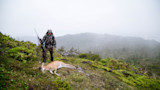 Hunter standing with rifle beside downed buck on mossy coastal ridge in fog