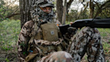 Hunter in camouflage sitting against tree, face masked, holding a shotgun