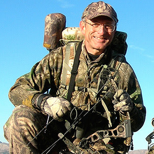 Man in camouflage holding a compound bow and backpack, smiling under blue sky