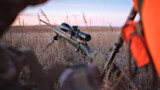 Rifle with scope on bipod in tall grass, blurred hunter wearing orange vest