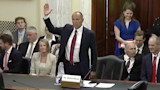 Man in suit raising right hand at a hearing; nameplate on desk reads "Mr. Michael Boren"