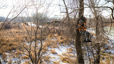 Hunter in camouflage and orange vest seated on a tree stand holding a bow