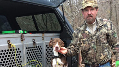 Hunter in camouflage holding brown-and-white dog beside pickup with kennels
