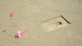Rectangular pit exposing ancient human footprint in sandy surface, pink flag and marker nearby
