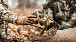 Hunter holding deer antlers, gloved hand labeled FIRSTLITE, two camouflaged hunters nearby