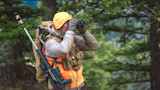 Hunter with orange beanie and vest scanning with binoculars, rifle slung