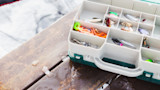 Open tackle box with fishing lures and orange soft plastics on a wet wooden dock