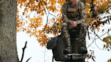 Hunter seated on a tree stand in camouflage with bow, arrows and orange autumn leaves