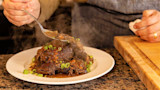 Steaming braised duck piled on white plate with sauce and sliced green onions, person spooning gravy