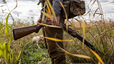 Hunter holding double-barrel shotgun in tall grass with yellow Labrador behind him