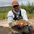 Man kneeling in shallow river holding a trout, wearing cap, sunglasses, and chest harness