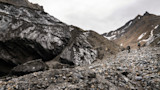 Two backpacked hikers walking across a rocky glacier valley under a cloudy sky