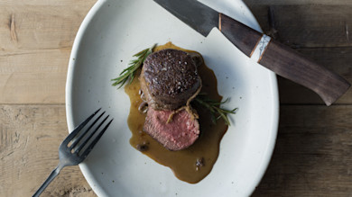 Porcini-crusted antelope filet with brown sauce and rosemary on white plate; knife and fork
