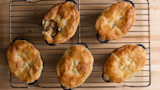 Six golden-brown pot pies on a cooling rack, one cut open revealing savory filling