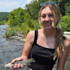 Woman holding a trout in a shallow stream with green trees and rocks behind her