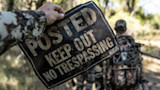Hand holding sign reading POSTED KEEP OUT NO TRESPASSING; hunter in camo blurred behind