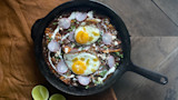 Cast-iron skillet of chilaquiles topped with two fried eggs, radish slices, crema, queso fresco, lime halves