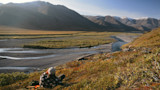 Man reclines on grassy hillside overlooking braided river and distant mountains
