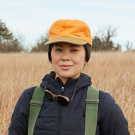 Woman wearing orange FILSON cap, black jacket and green suspenders in grassy field