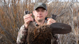 Steven Rinella holding a beaver by its hind feet, wearing a cap labeled VORTEX