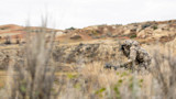 Hunter in full camouflage kneeling with rangefinder and crossbow on grassy bluff in badlands
