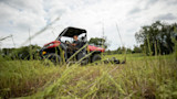 Man driving red side-by-side UTV across meadow mowing tall grass
