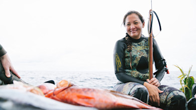 Woman in camo wetsuit holding a speargun on a boat beside harvested fish