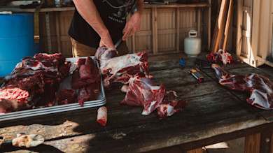 Butchered venison on wooden table as a person carves meat; T-shirt reads 'MEATEATER'