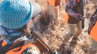 Hunter wearing blue knit hat and gray cap, shotgun over shoulder; other hunters in orange vests in dry grass