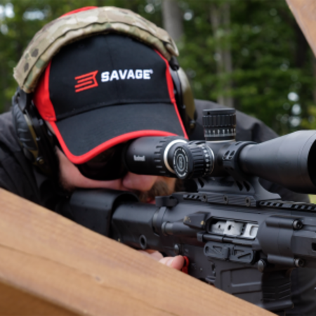 Hunter aiming a rifle through a scope, wearing ear protection and a cap reading "SAVAGE"