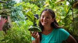 Smiling woman holding a leaf-wrapped lau lau parcel in a lush garden
