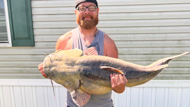 Man with glasses and red beard holding huge catfish in front of vinyl house siding