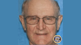 Older man wearing glasses, close-up portrait on blue background with TBI seal