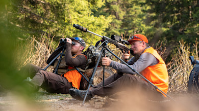 Two hunters in orange vests, one using binoculars, one aiming a rifle on a tripod in forest