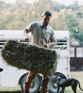 Man lifting a hay bale beside a white trailer with a black dog nearby