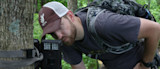 Man inspecting a trail camera strapped to a tree in a leafy forest