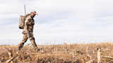 Hunter carrying shotgun over shoulder with backpack walking across harvested cornfield wearing camo