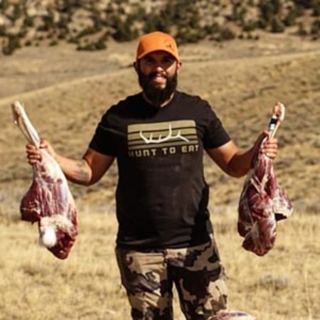 Man in orange cap and HUNT TO EAT shirt holding two skinned animal carcasses in a field