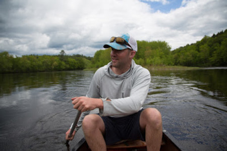 Man paddling canoe on river with treed shoreline and cloudy sky