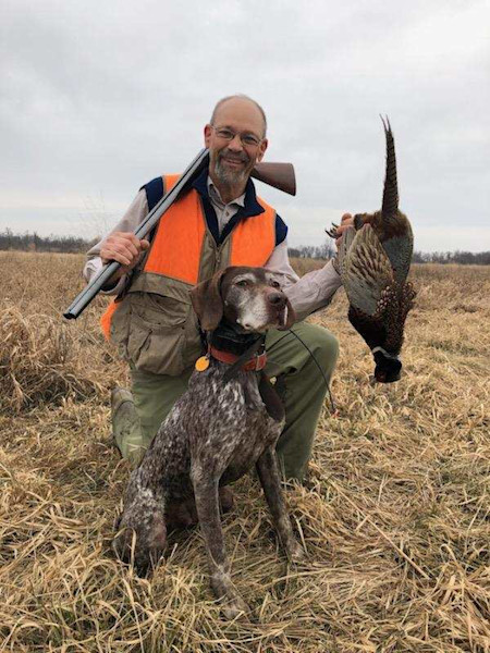Man kneeling in field with shotgun and a pheasant, German shorthaired pointer sitting beside him